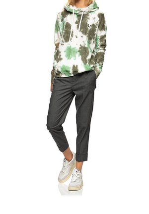 Osoba ubrana w zielono-brązową bluzę z kapturem w stylu tie-dye, szare spodnie z mankietami i białe trampki, stojąca z rękami w kieszeniach.
