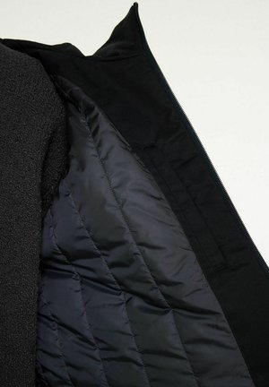 Svart jacka med en vadderad nylonfoder och en mjuk fleecepanel inuti, delvis uppfälld för att visa interiörens detaljer.