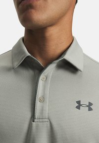 Polo gris clair à col avec une surface texturée, doté d'une patte à trois boutons et d'un logo de marque gris foncé sur la poitrine.