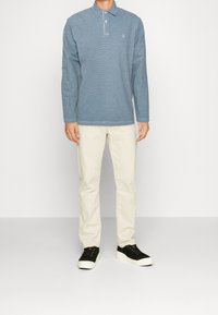 Langärmliges blaues Poloshirt mit Kragen und kleinem Logo, kombiniert mit hellbeigen Hosen und schwarzen Turnschuhen mit weißen Sohlen.