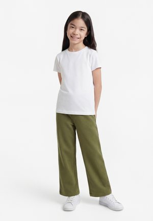 Noor tüdruk pikkade tumedate juustega kannab valget lühikese varrukaga T-särki, oliivrohelisi laiade jalgadega pükse ja valgeid tossusid, naeratab ja seisab.