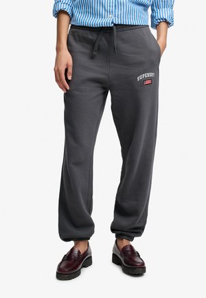 Model iført mørkegrå Superdry joggingbukser med elastiske manchetter og snøre i taljen, kombineret med bordeaux loafers og en blåstribet skjorte.