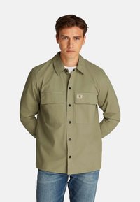 Chaqueta casual verde oliva con dos bolsillos en el pecho, cierre de botón y un cuello. Hecha de tela texturizada, combinada con jeans azules.