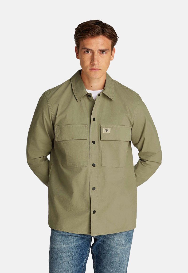 Chaqueta casual verde oliva con dos bolsillos en el pecho, cierre de botón y un cuello. Hecha de tela texturizada, combinada con jeans azules.