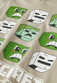 Camiseta beige con caras de estilo caricaturesco en bloques, con diversas expresiones. Los colores incluyen verde y negro con rasgos faciales y contornos distintivos.