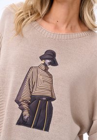 Beżowy sweter dzianinowy z graficznym nadrukiem postaci w pasiastym stroju z żółtymi akcentami i czarnym kapeluszem. Teksturowana konstrukcja materiału.