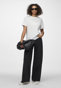 Weißes T-Shirt, weite schwarze Jeans und schwarze übergroße Handtasche. Das Model trägt schwarze Sonnenbrillen; der Hintergrund ist rein weiß.