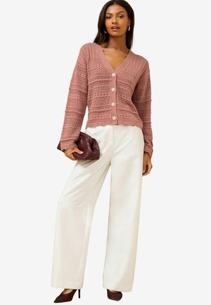Häkelstrickjacke in zartem Rosé mit V-Ausschnitt, gewelltem Saum und großen Knöpfen; kombiniert mit weißen, weiten Hosen und einer burgunderfarbenen Clutch.