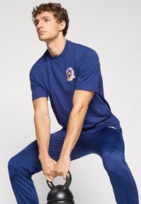 Mørkeblå atletisk T-shirt med korte ærmer, der har et farverigt logo på brystet. Modellen holder en sort kettlebell.
