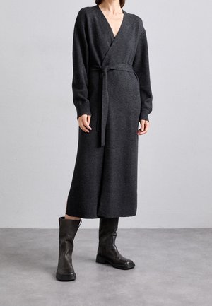 Kobieta ubrana w długą, ciemnoszarą dzianinową sukienkę z wiązanym paskiem i czarne skórzane kozaki sięgające połowy łydki, stojąca na szarej podłodze.