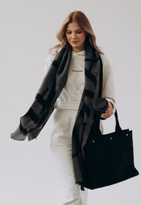 Zwarte tote bag met reliëftekst, grijze sjaal met gedurfde print, witte hoodie en crèmekleurige broek; houdt een zwart koffiemok vast.