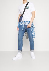 Bílý tričko s logem Levi's, modrá vázaná flanelová košile kolem pasu, modré skinny džíny a bílé tenisky. Minimalistický design.
