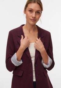 Bordeauxroter Blazer mit gekerbtem Revers, zwei Vorderfächern und grauen gestreiften Manschetten, getragen über einem weißen Oberteil, mit glatter Textur.