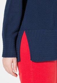 Hand neben einem marineblauen strukturierten Pullover mit seitlichem Schlitz, der über roten Hosen getragen wird.