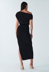 Czarna, dopasowana sukienka maxi z jednym odsłoniętym ramieniem, miękka tkanina, rozcięcie z boku oraz gładkie, eleganckie wykończenie. Złote sandały na obcasie.