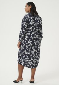 Floral wrap jurk in marineblauw met witte en lichtblauwe bloemen. Heeft lange mouwen, een strik in de taille en een lengte tot halverwege de kuit.