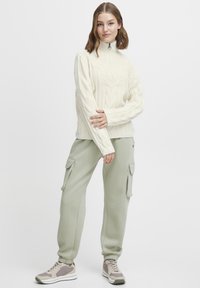Pull en laine beige à torsades avec col montant, associé à un pantalon cargo vert clair avec poches latérales, et des baskets gris-blanc.