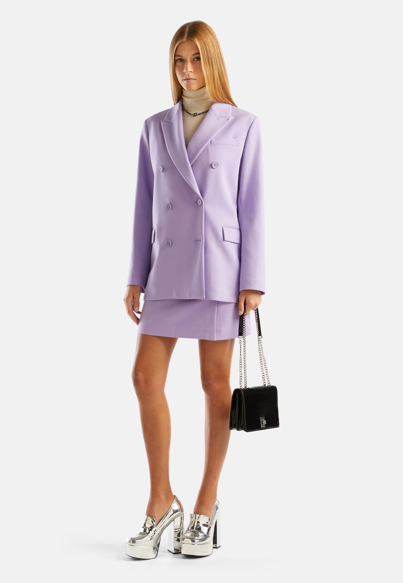 Lavenderfarbenes doppelt geknöpftes Blazerjackett mit passendem Minirock, kombiniert mit schimmernden silbernen Plateau-Schuhen und einer kleinen schwarzen Handtasche mit Kettenriemen.
