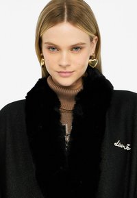 Giovane donna con capelli biondi che indossa orecchini a forma di cuore dorati, un dolcevita marrone a maglia e un cappotto nero con colletto di pelliccia e spilla.
