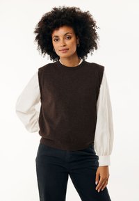Un maglione estivo marrone lavorato a maglia senza maniche sovrapposto a una camicia bianca a maniche lunghe con texture, abbinato a jeans neri con bottoni laterali.