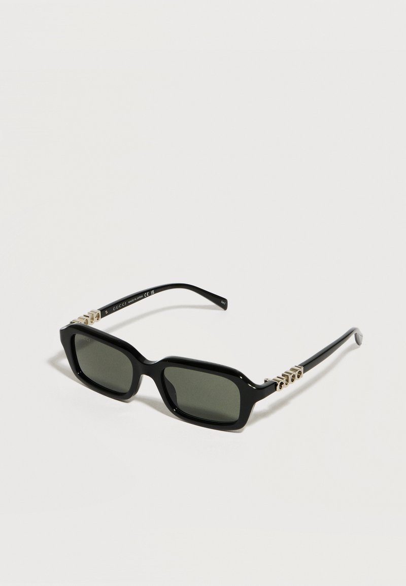 Lunettes de soleil rectangulaires noires avec verres teintés verts et accents métalliques du logo Gucci sur les branches, présentées sur un fond blanc.