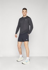 Camisola desportiva de manga longa preta combinada com calções azul-escuros, apresentando um logótipo. O modelo também usa sapatilhas brancas e azuis com meias brancas.