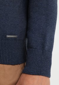 Tummansininen neulepusero, jossa on ribbireunat, teksturoitu kangas ja pieni hopeinen etiketti, jossa lukee "PIER ONE" sivussa.
