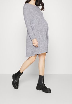 Femme enceinte en robe grise ample à manches longues portant des bottines noires épaisses, marchant devant un fond blanc uni.