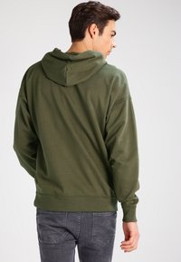 Felpa verde in cotone con cappuccio regolabile, polsini a coste, vestibilità rilassata e tasca a marsupio, abbinata a jeans grigi.