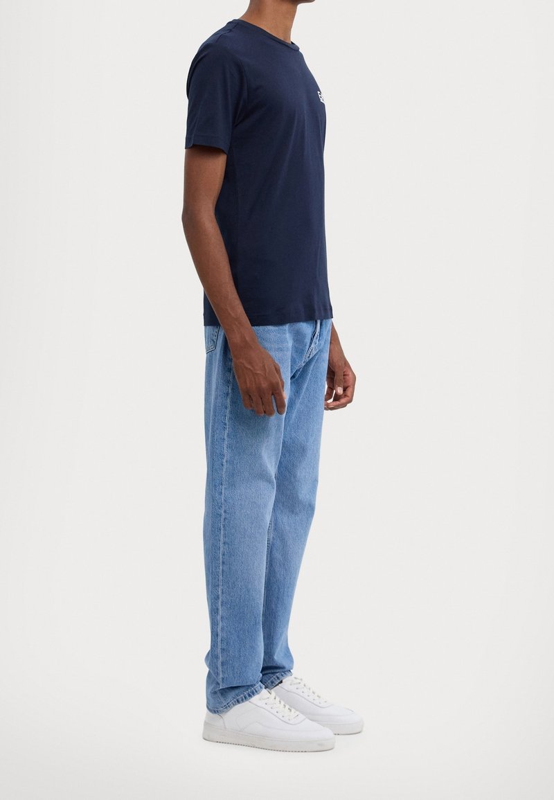 Mörkblå bomullströja med rund hals, kombinerad med ljusblå denimjeans och vita sneakers, visad från sidan mot en enfärgad bakgrund.