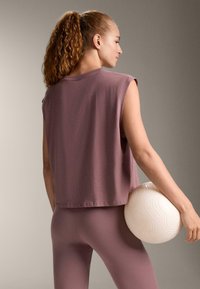 Top fără mâneci, cu croială lejeră, din material mauve estompat, asortat cu colanți strâmți, în aceeași nuanță. Modelul ține o minge de exerciții texturată, de culoare alb murdar.