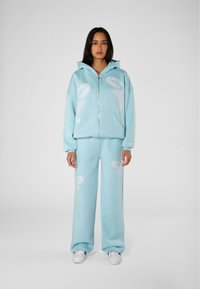 Felpa azzurra chiara e pantaloni coordinati realizzati in morbido tessuto; presenta grandi motivi astratti bianchi e una chiusura con zip. Sneakers bianche completano il look.