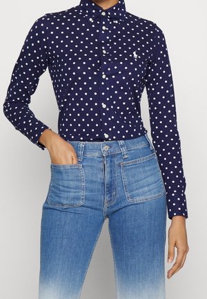 Person iført en mørkeblå skjorte med lange ærmer og hvide polkaprikker samt lyseblå jeans med høj talje, hånd i lomme.