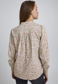 Langærmet blomsterbluse med en lys beige baggrund, prydet med pink og grønne blomster, med rynkede detaljer ved ærmerne og en knaplukning bagpå.