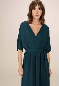 Teal, wikkelstijl jurk met een diepe V-hals, korte mouwen en een gestrikte taille. Gemaakt van een lichte, gestructureerde stof die naar de vloer stroomt.