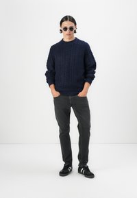 Jersey de punto azul marino con un patrón de trenzas texturizado, jeans grises y zapatillas negras con acentos blancos. El modelo lleva gafas de sol redondas.