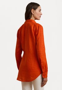 Orange linen shirt featuring a button-down collar, long sleeves with button cuffs, a curved hem, and a textured finish.