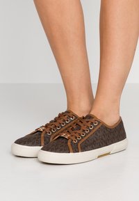 Braune Canvas-Sneaker mit Muster, braunen Lederakzenten und weißen Gummisohlen. Verfügt über eine runde Zehenpartie und eine Schnürung vorne.