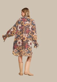 Blomstrete gjennomsiktig kjole med gule og røde blomster, lange ermer med rynkede mansjetter, og løs passform, kombinert med beige regnstøvler.