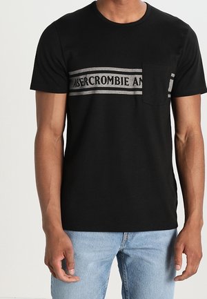 Czarna koszulka z krótkim rękawem z szarym poziomym paskiem i napisem "Abercrombie and Fitch", noszona przez osobę w jasnoniebieskich dżinsach.