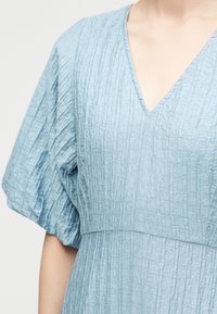 Φόρεμα από υφή ελαφρώς γαλάζιου υφάσματος με βαθύ V-λαιμόκοψη και κοντά φουσκωτά μανίκια, που εμφανίζεται σε ένα πρόσωπο από τον λαιμό έως τη μέση.