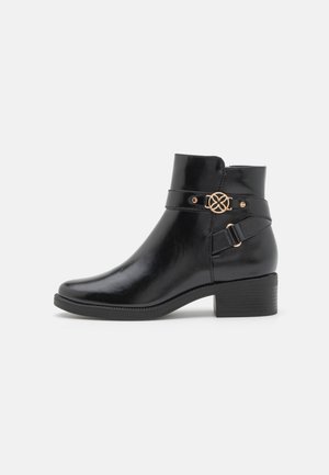 Botte de cheville en cuir noir avec une finition brillante, présentant un accent décoratif en or et un détail de sangle autour de la cheville, talon carré.