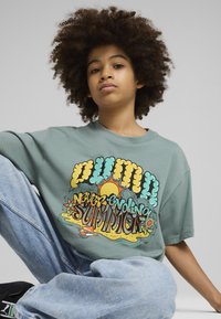 Camiseta de algodón verde con un diseño gráfico colorido que incluye el texto "nunca evolucionando verano", combinada con unos pantalones de mezclilla azul claro.