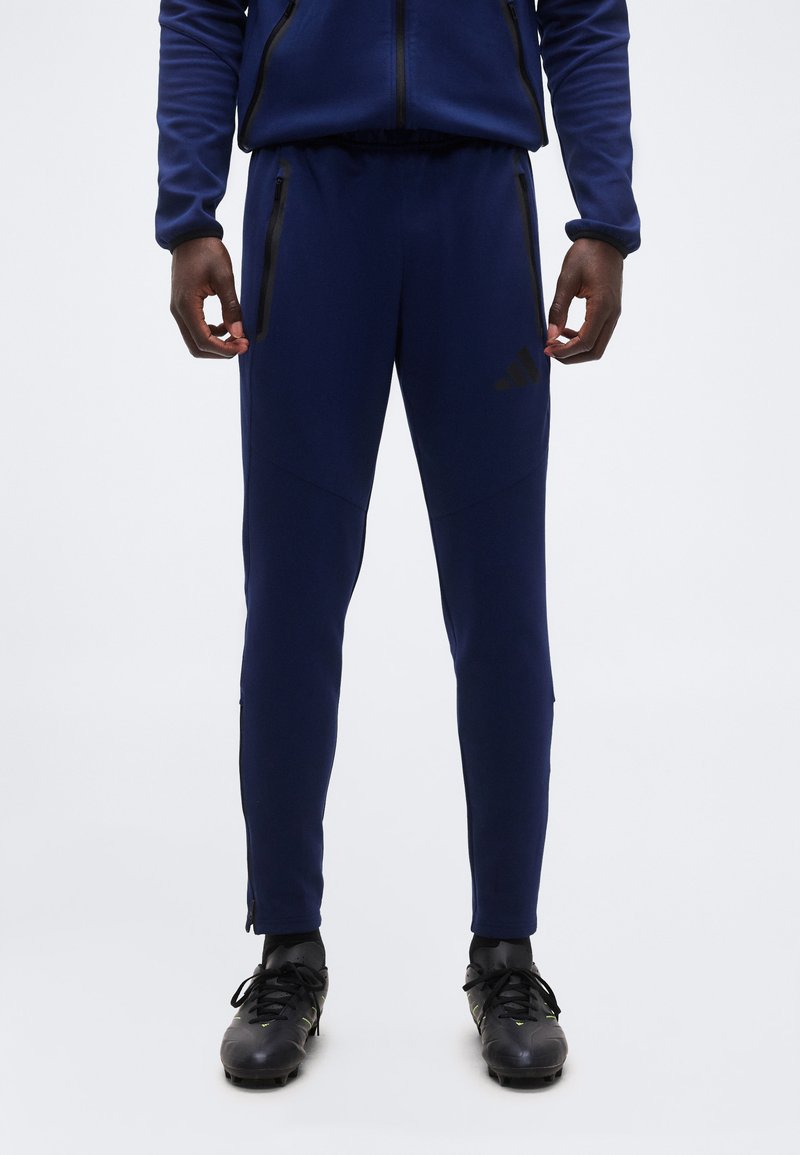 Άνδρας που φοράει ναυτικό μπλε παντελόνι τζόγκινγκ και μαύρα ποδοσφαιρικά παπούτσια, στέκεται με τα χέρια χαλαρά δίπλα στο σώμα σε λευκό φόντο.