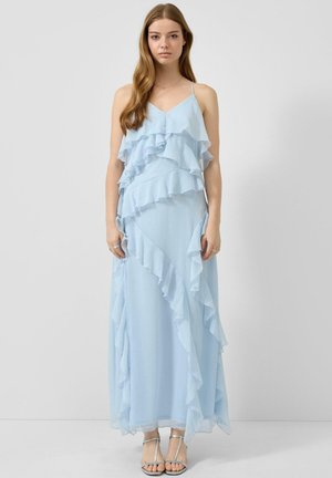 Vrouw draagt een lichtblauwe mouwloze maxijurk met gelaagde ruches en dunne schouderbandjes, gecombineerd met zilveren open-toe hakken.