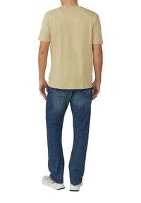 Beige katoenen t-shirt met korte mouwen, ronde hals, gedragen met blauwe spijkerbroek en lichte sneakers. Achteraanzicht.