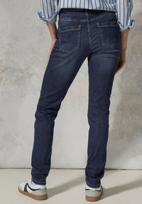 Dark blue skinny jeans with back pockets, made of denim fabric, featuring a smooth texture and a tailored fit. Paired with white sneakers.