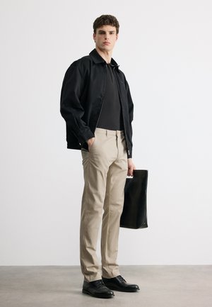 Mladý muž oblečený v čiernej bunde, čiernom tričku, béžových nohaviciach a čiernych topánkach, držiaci veľkú čiernu koženú tašku v neutrálnom štúdiovom prostredí.