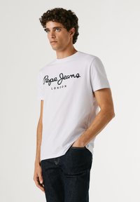 Nuori mies, jolla on valkoinen Pepe Jeans London -t-paita ja tummat farkut, seisoo yhdellä kädellä taskussa yksiväristä taustaa vasten.