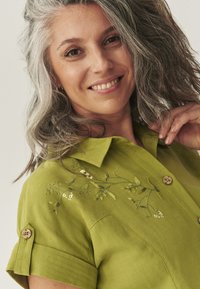 Camicetta verde a maniche corte con colletto, caratterizzata da ricami floreali bianchi e verdi. Bottoni in legno abbelliscono i polsini e la parte frontale.